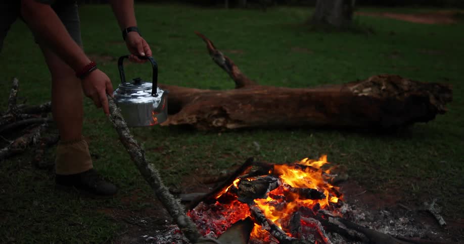 A man stoking a campfire with a log while holding a kettle