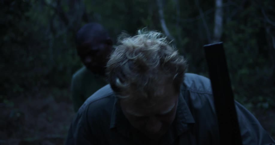 A man carrying a firearm over his shoulder leads another man through a forest before kneeling to examine the ground