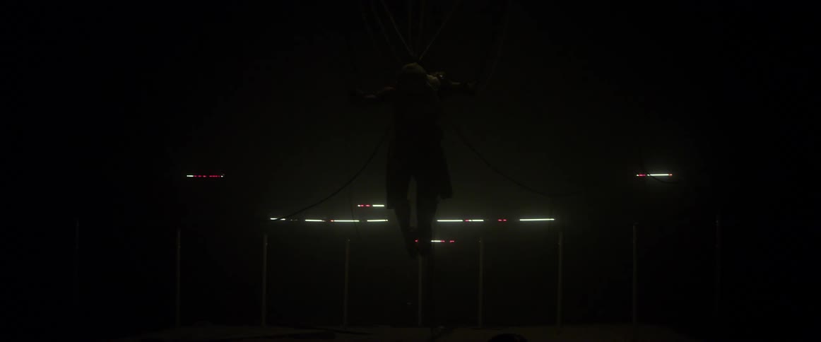 A person is suspended by cables in a dark room with flashing light