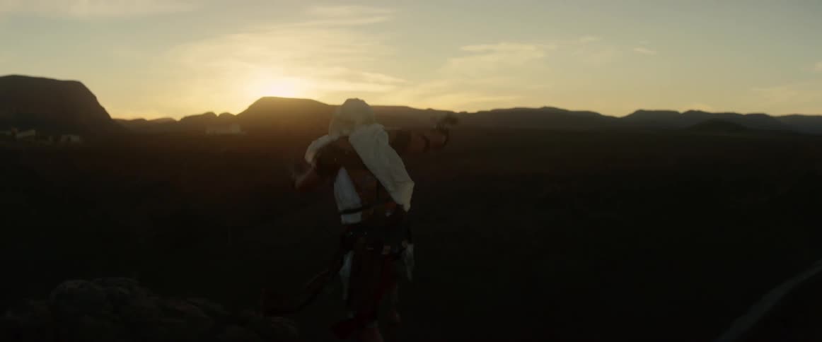 A man wearing a white headscarf jumps high as overlooks a canyon in the distance