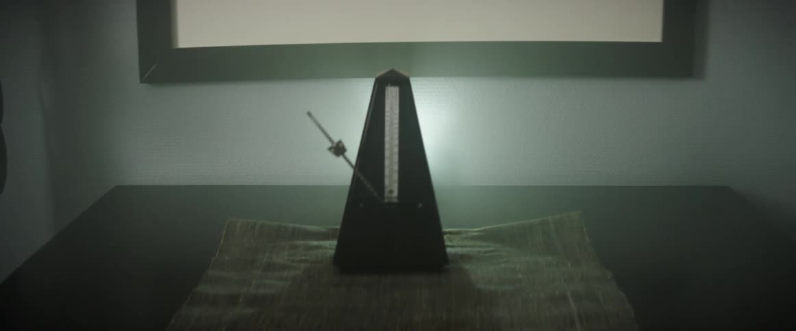 Metronome arm swings on wooden table in dim room near picture on wall