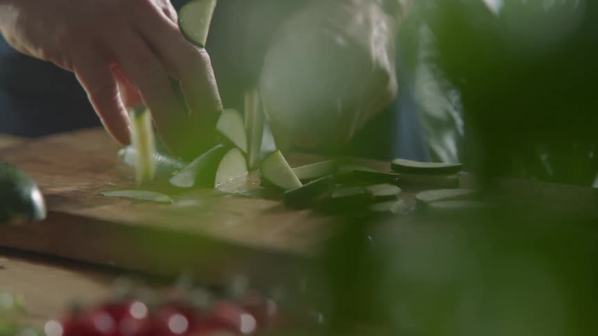 Man slices a cucumber thinly with a sharp knife on a cutting board in the kitchen