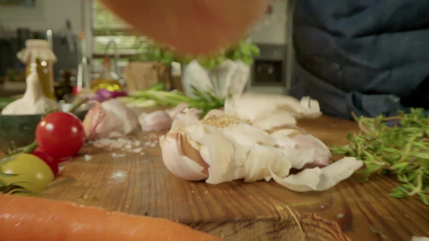 A man places a garlic bulb on a cutting board full of other bright colorful vegetablesand smashes it