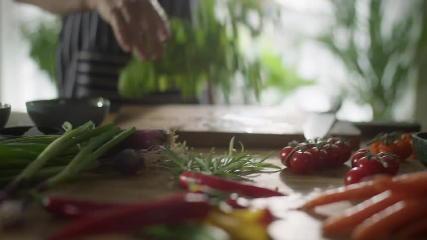Assorted vegetables sit on a table while a woman drops some lettuce