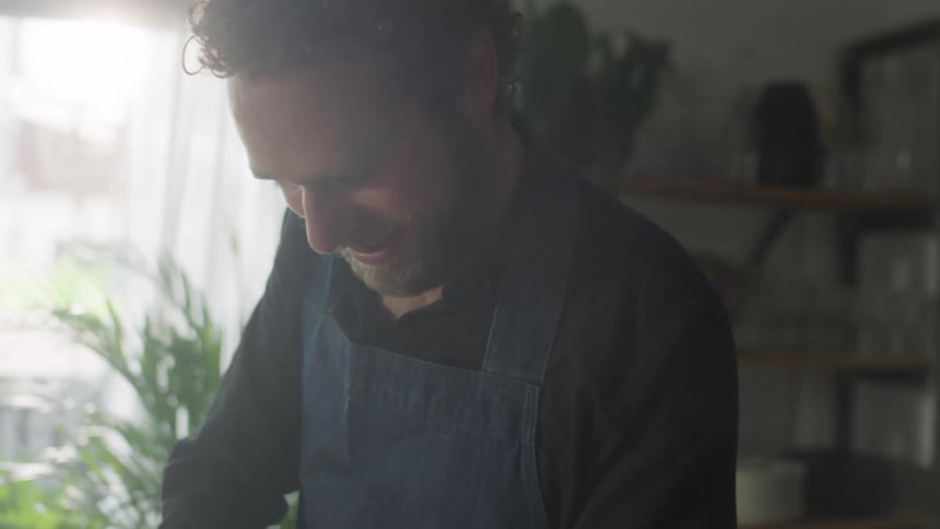 Man with a beard and blue apron cuts vegetables with a knife on a cutting board in the kitchen