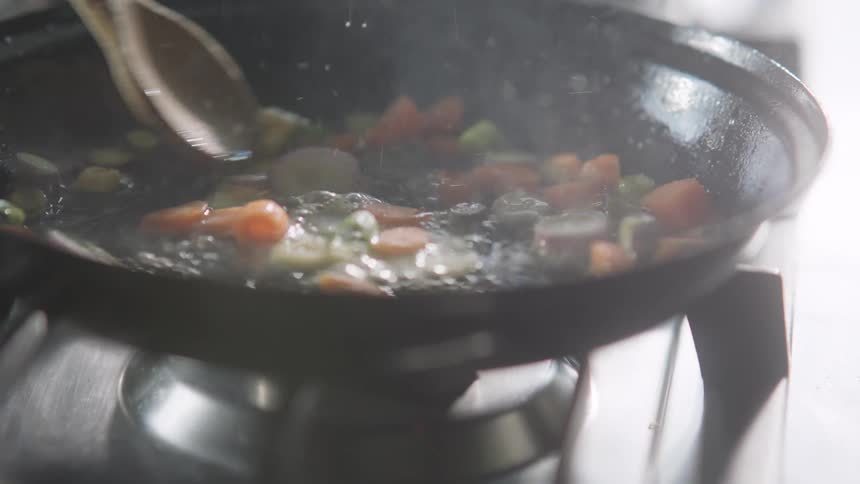 A person uses a wooden spoon to stir vegetables being fried with oil in a black skillet