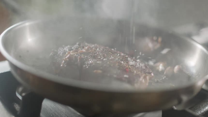 A person sprinkles salt and seasoning on a piece of meat cooking in a pan