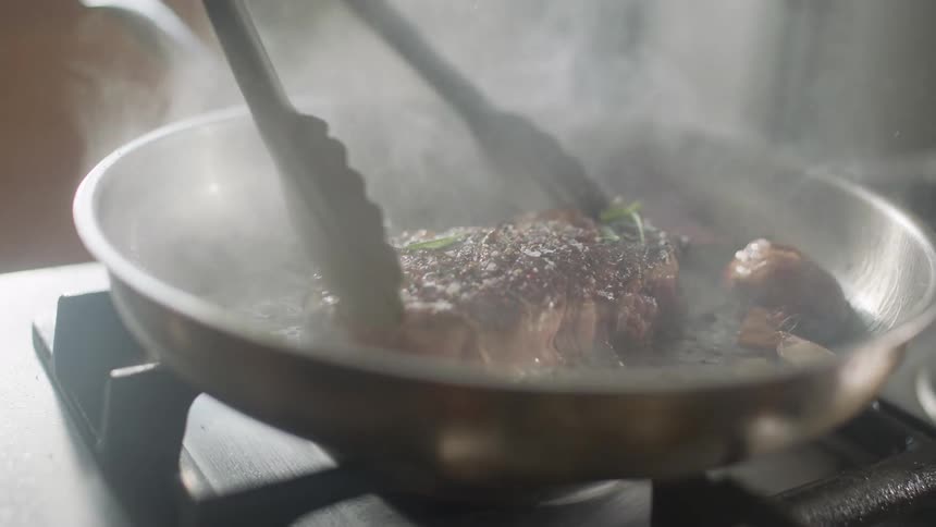 A pair of tongs move a piece of steak in a frying pan