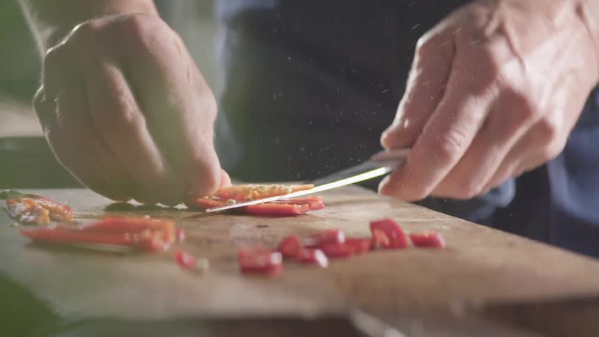 Cook with a dark blue apron cuts a red hot chili pepper with a knife on a cutting board in the kitchen