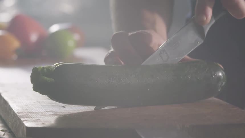 Cook with a black shirt cuts a zucchini in half with a knife on a cutting board in the kitchen