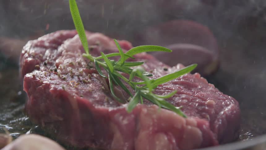 Pepper flakes fall onto steak topped with rosemary sitting among oil in pan