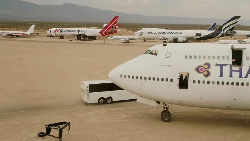 An airport in the desert with a bus driving by airplanes with mountains and the sky in the background