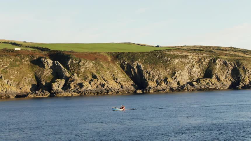 A person on a boat on a lake close to a rocky coast with a field and the sky in the background
