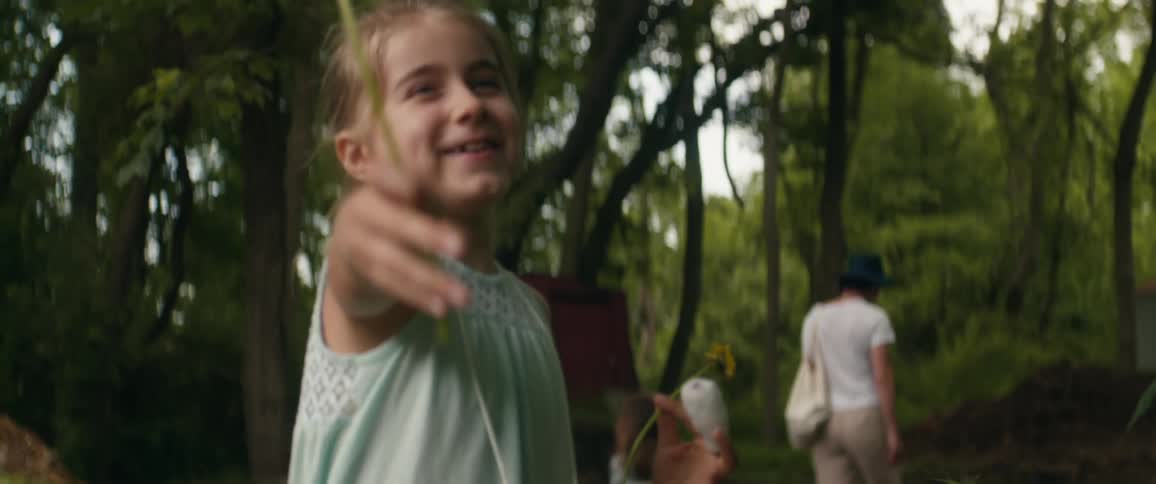 A smiling little girl hands a dandelion to a person out of frame