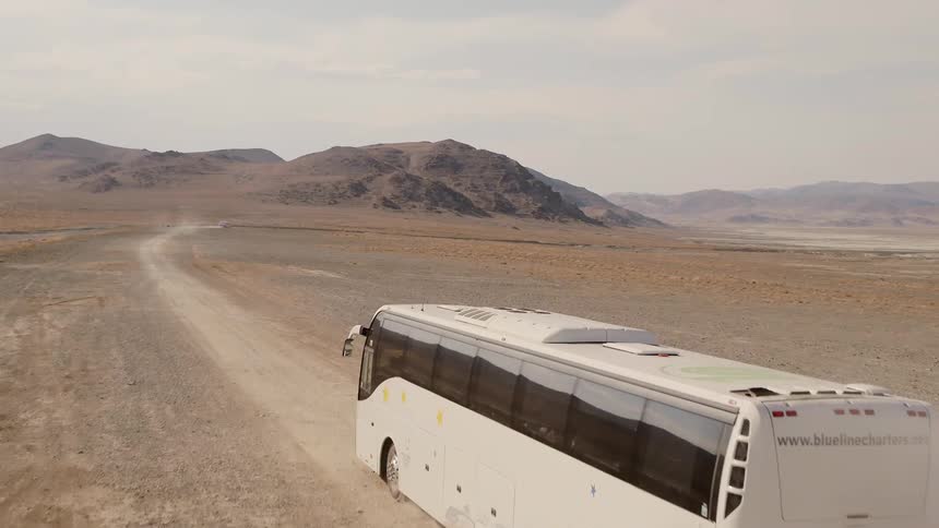 Bus drives along dirt road through desert near low mountains kicking up dust