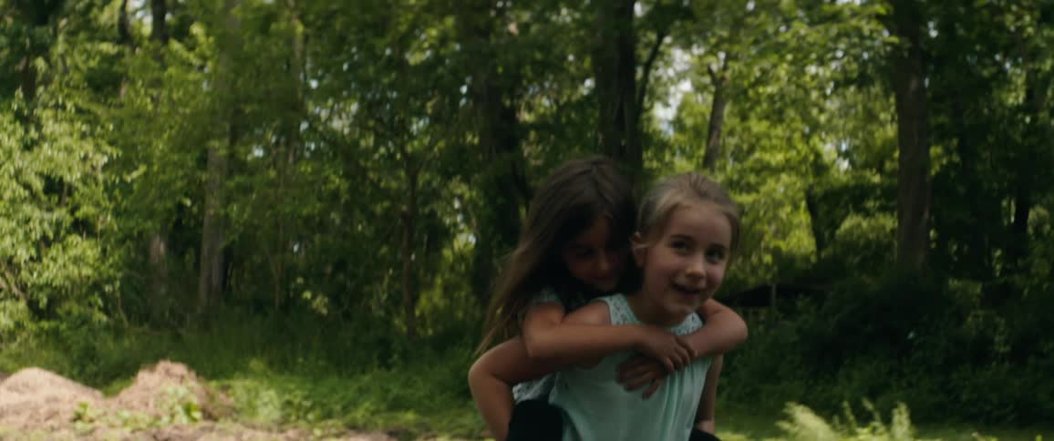 A little girl gives her sister a piggy back ride through a forest