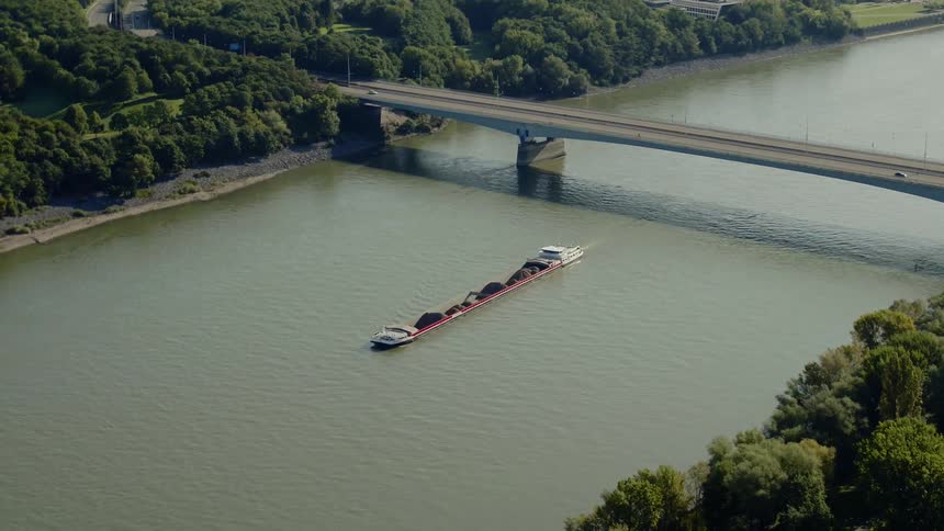 A shipping boat sails on a small river by a bridge and trees