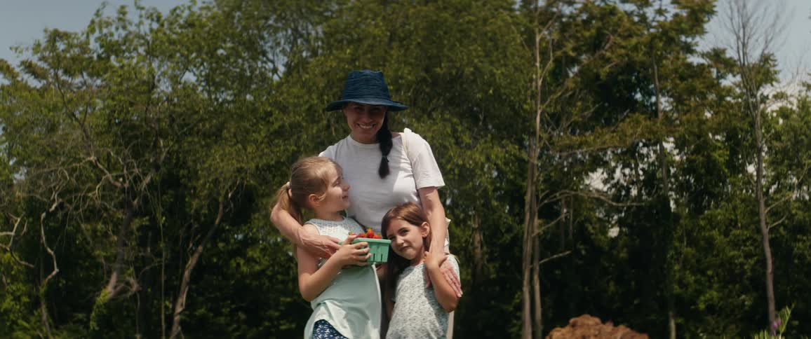 A woman holds on to her two young daughters outside while they are holding fresh fruit