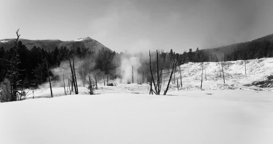 Smoke rises from snow covered ground near conifer forest backed by mountains