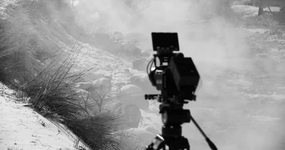 Photography camera mounted on a tripod in front of a steaming river and a deer grazing the snow covered grass