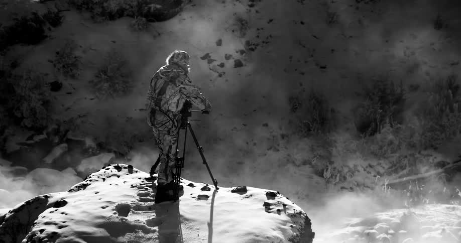 A man stands on a snowy rock and films an area with steam coming out of it