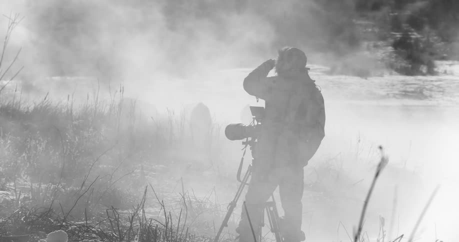 Man in winter gear stands next to a camera on a tripod in the mist