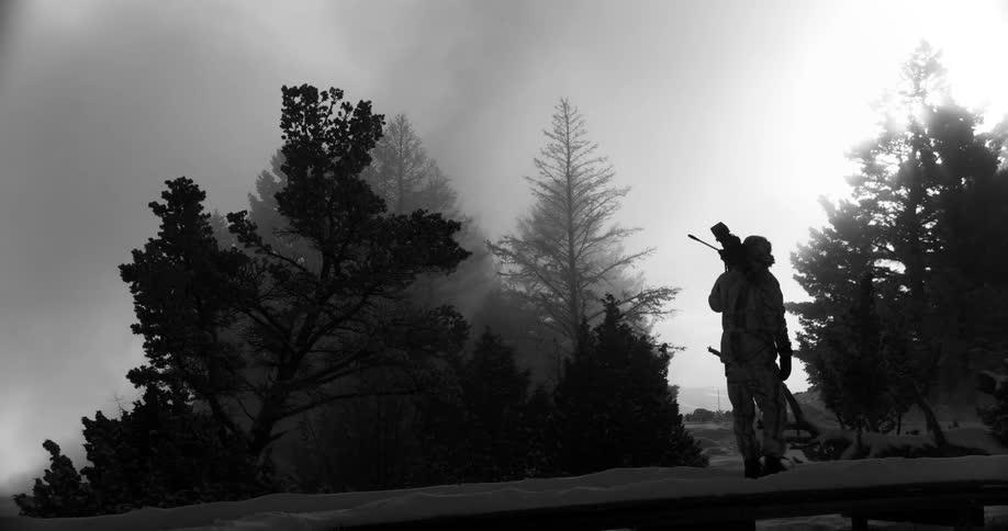 A man carries a film camera on tripod across a snow covered bridge pausing to look at the smoke billowing from behind the trees