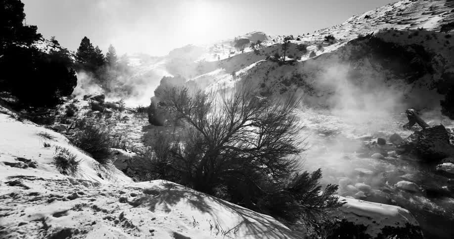 Steam rises from a stream running through a hilly snow covered area