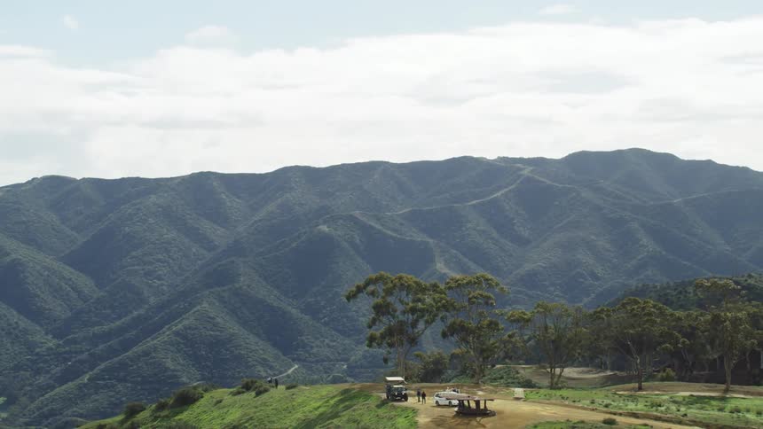 People standing next to vehicles with trees hills sky and clouds in the background