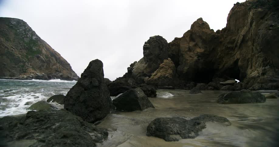 Waves crash on a beach that lies below rocky cliffs
