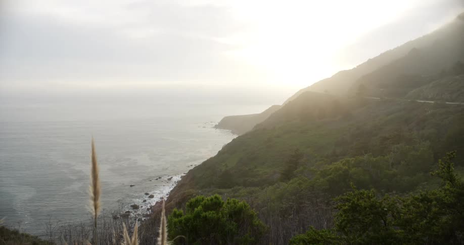 Mist covers green mountain bordering water as sun shines on horizon