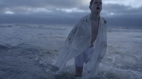A man removing a white sheet from his head as he wades out of a rough ocean on a cloudy day