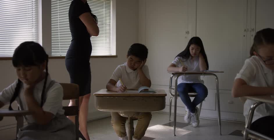 A female teacher walks around the desks of her elementary students writing