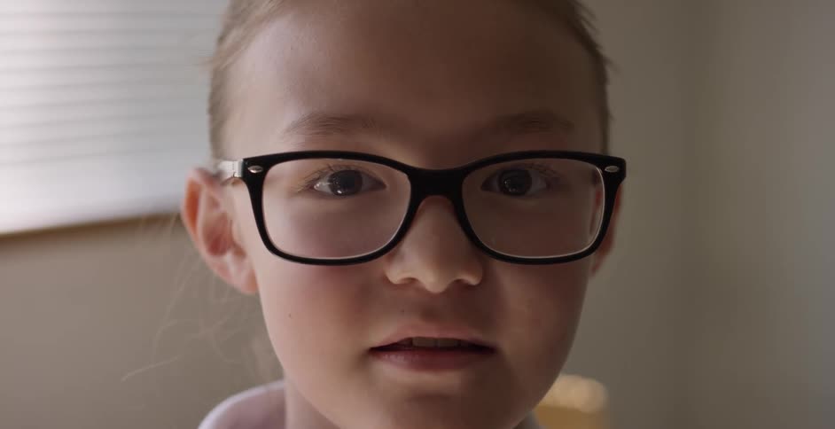 Girl wearing dark rimmed eyeglasses talks in front of window with blinds closed