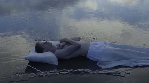 A white sheet is slowly pulled off a person lying on the wet sand