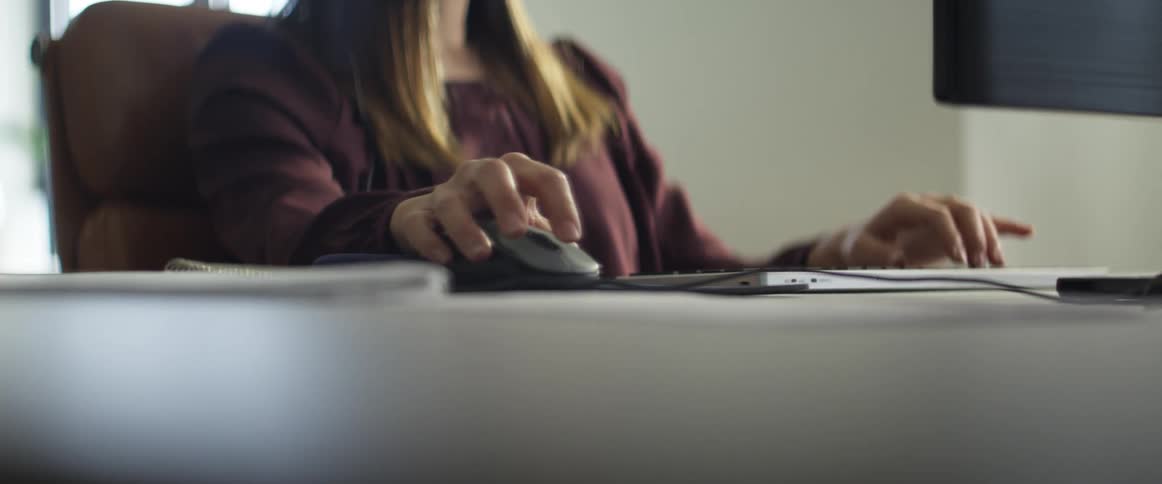 Woman with a long sleeved blouse sitting at the desk uses a mouse and types on a computer keyboard