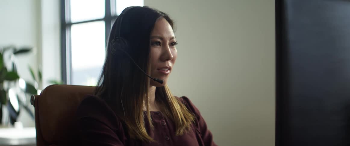 A woman sits at an office desk in front of a computer talking on a headset