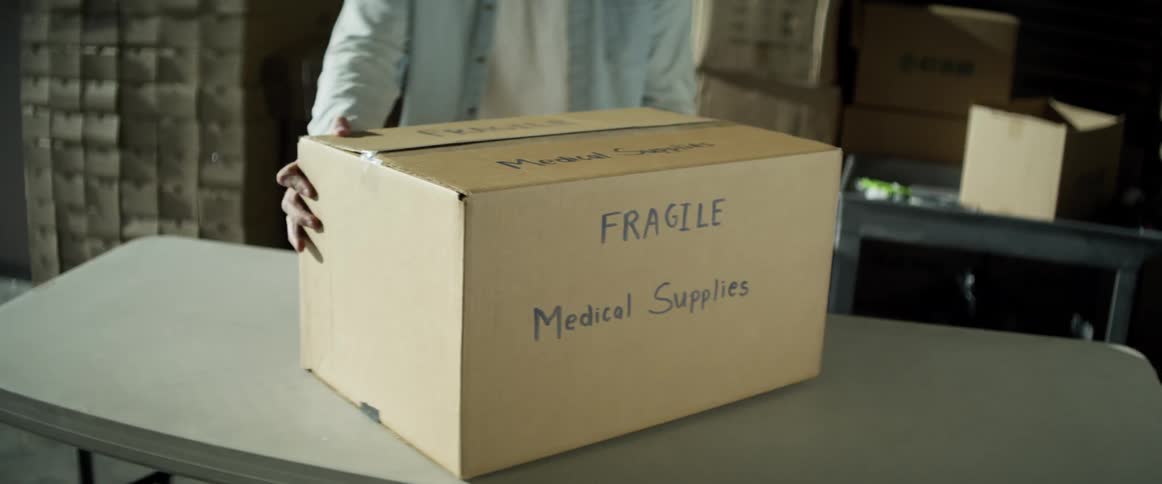 Man with a light blue shirt and jeans carries a box of medical supply and puts it on the table