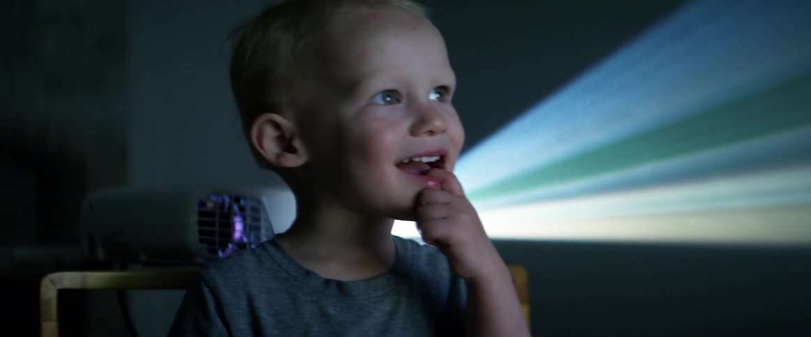 A woman tickles a little boy in the their house as a projector plays in the background