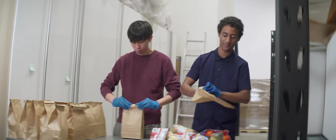 Two men pack food into paper bags for lunches