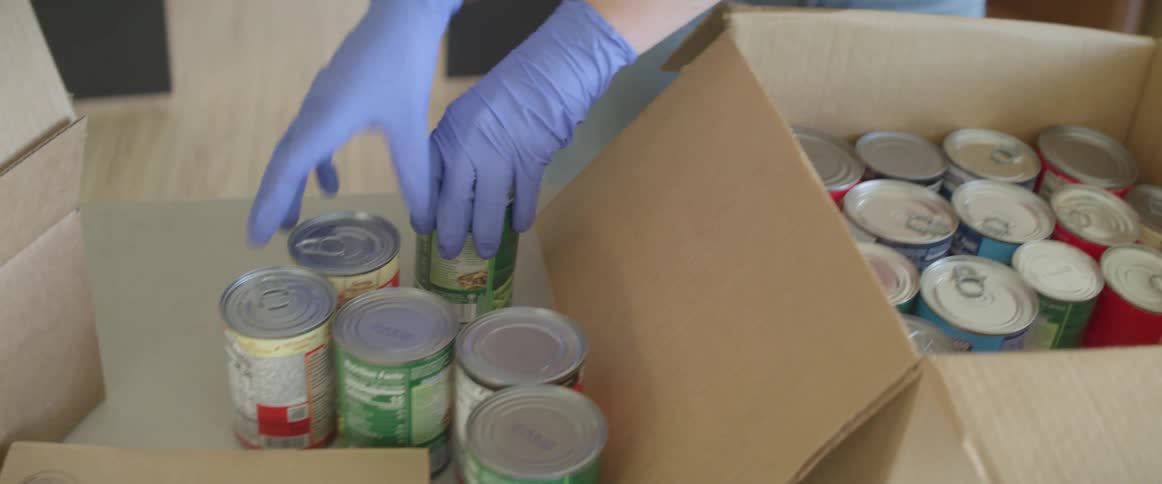 Person wearing latex gloves fills a cardboard box with canned food in a warehouse