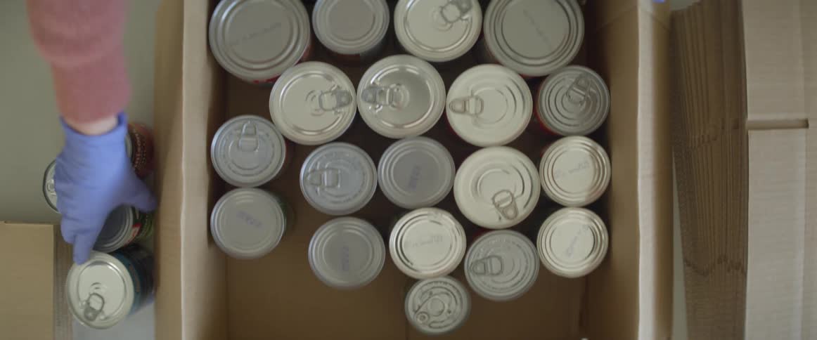 A person wearing gloves places canned food into a cardboard box