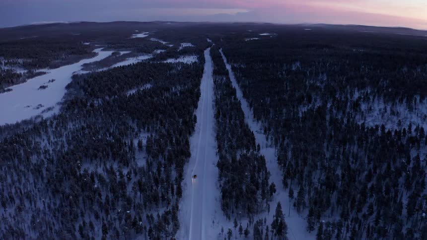 A vehicle drives down a snow covered road flanked by trees in a forest
