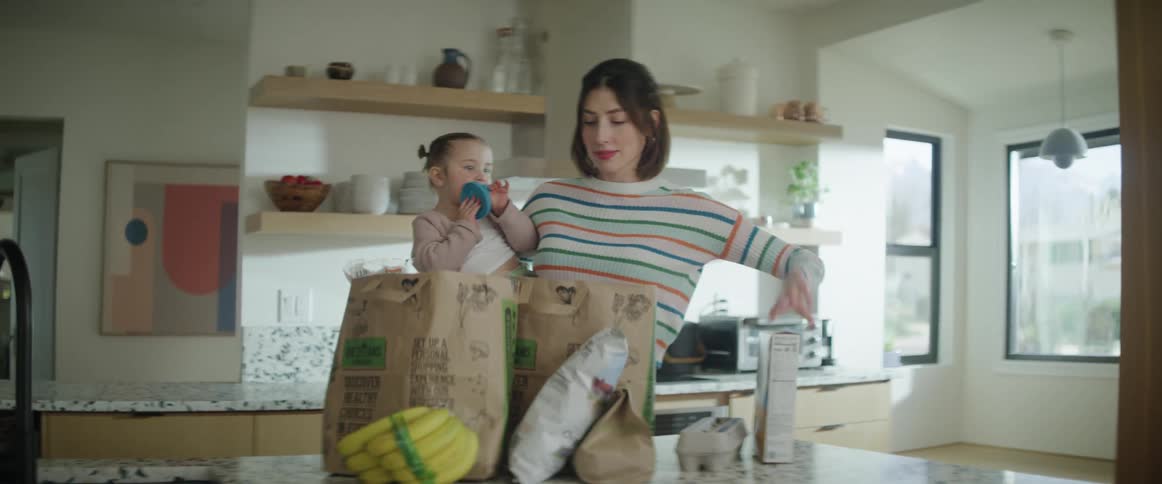 Young mother holding toddler unloads groceries from brown paper bags in kitchen