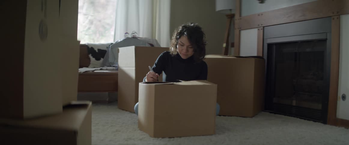 Woman sits on living room floor writing on moving boxes