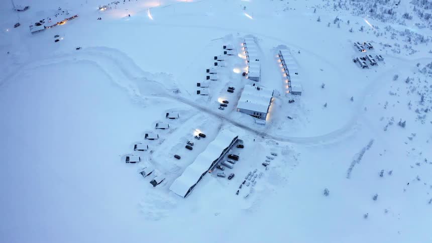 Vehicles surrounding buildings and nearby woods are all covered in snow