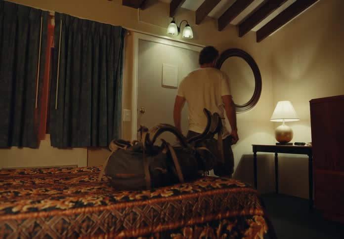 A man walks in to a hotel room throws his bag on the bed and goes and looks in a mirror