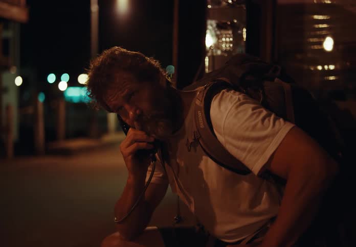 A man sits on a city street and talks on a payphone
