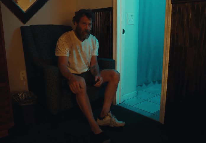 A man sits on a chair outside of his bathroom and takes off one shoe angrily