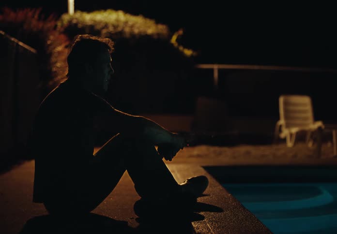 A man sits beside a pool and lights a cigarette at night